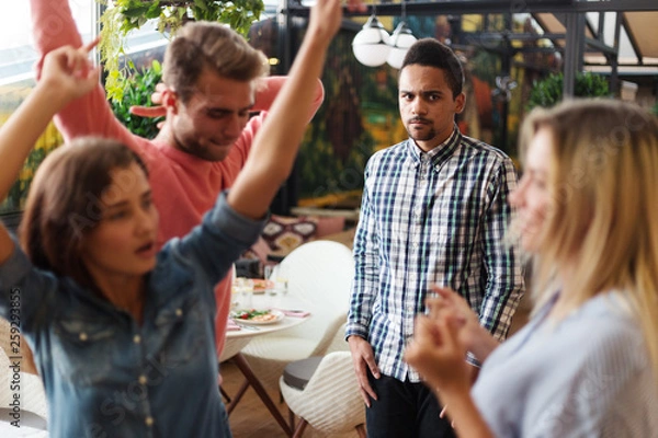 Fototapeta Sad jealous boyfriend standing aside and watching his girlfriend having fun and dancing with friends at party
