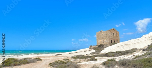 Fototapeta empty white beach with old ruins of abandoned stone house on the rocks and summer blue sky and sea in background in Sicily Italy