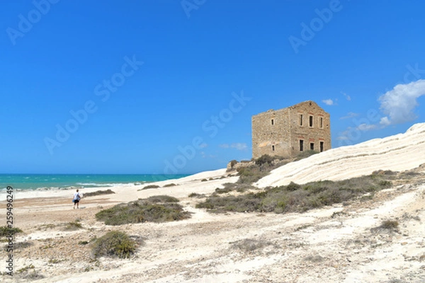Fototapeta empty white beach with old ruins of abandoned stone house on the rocks and summer blue sky and sea in background in Sicily Italy