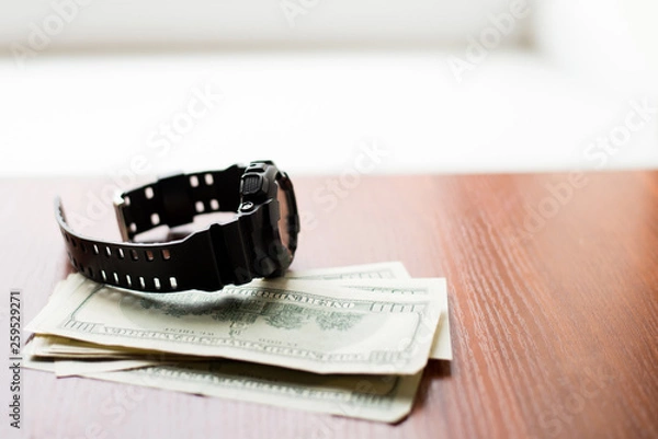 Fototapeta Time is money. One hundred dollars and black watch on wooden table.