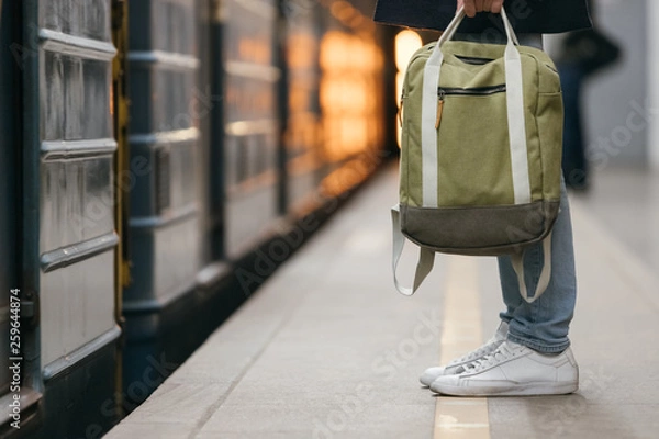 Obraz Close up photo of male waiting subway train on platform. Well-dressed handsome man in white sneakers and jeans with backpack in hand standing on station. Modern citizen of big city. Lifestyle concept.