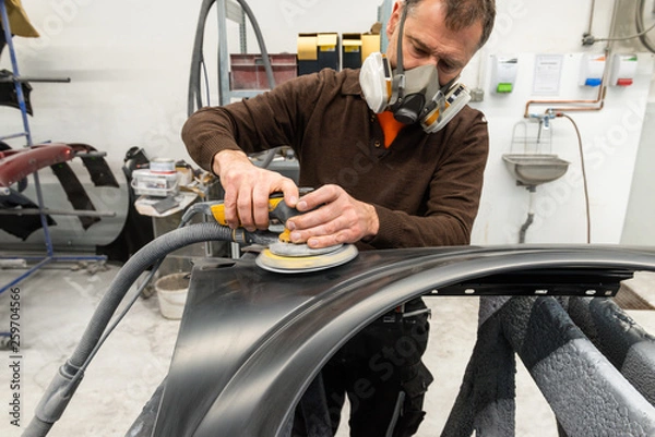 Fototapeta Professioneller Autolackierer schleift ein Autoteil mit der Schleifmaschine vor in einer Werkstatt - Serie Autowerkstatt