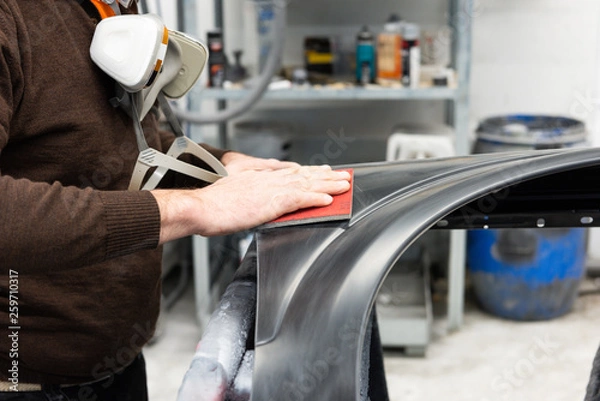 Fototapeta Professioneller Autolackierer schleift ein Autoteil mit Schmirgelpapier vor bei in einer Werkstatt - Serie Autowerkstatt.