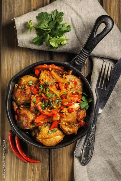 Fototapeta Fried chicken in spicy sauce with vegetables. Cooked in a cast iron pan. The pan is on a dark wooden background. Next is a natural linen napkin. Top view. Vertical.