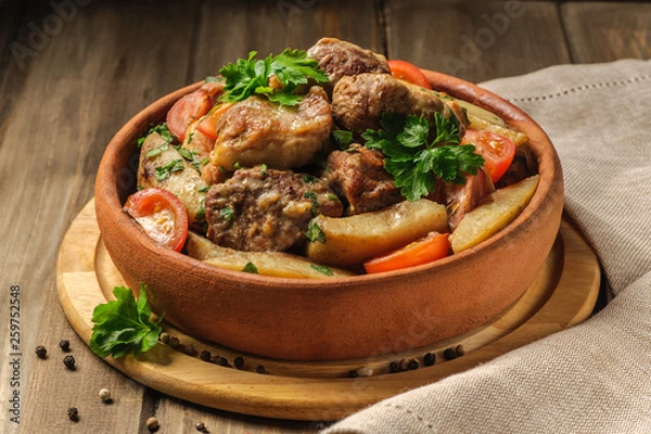 Fototapeta The traditional pork and potato dish of Georgian cuisine on earthenware ketsi. Ojahuri with tomatoes, hot peppers and fresh herbs. Dark background and natural napkin. Horizontal view.