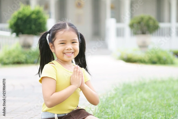 Fototapeta Asian child smiling hand in hand or kid girl pay obeisance and pray or hello welcome and thank you with wear yellow shirt in morning at hotel and church or temple for peace on lawn and receptionist