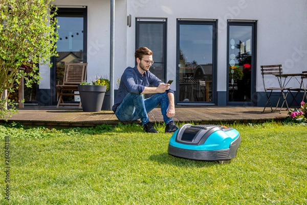 Fototapeta junger Mann sitzt mit Handy hinter Roboterrasenmäher, Garten