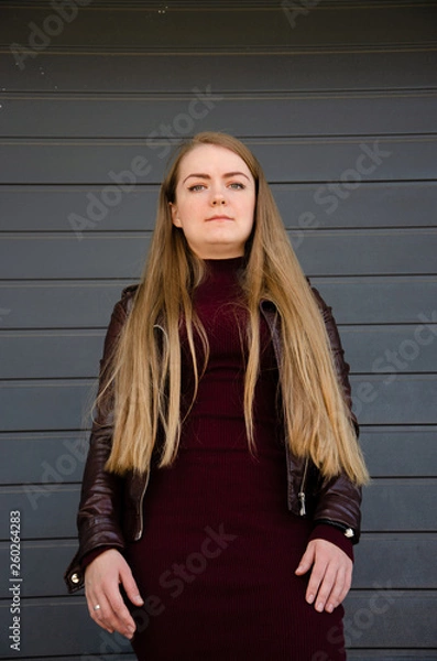 Obraz Portrait of a beautiful girl in a dark red dress knee-deep and in a dark leather jacket in the street in the afternoon