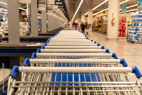 Fototapeta A lot of supermarket carts handles with shallow depth of field with a part of the market view blurred