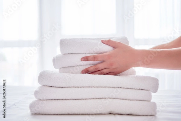 Obraz Woman putting stack of fresh white bath clean towels on bed sheet, copy space. Close up hands of hotel maid with towels. Room service