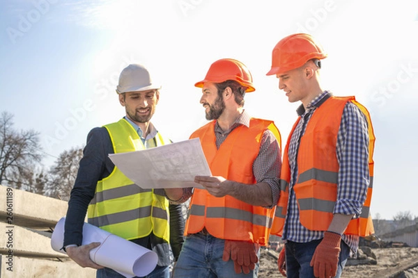 Fototapeta male builders workers engineers at a construction site looking drawings in helmets