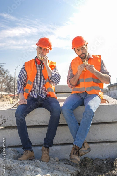 Fototapeta two workers in helmets lunch, rest, sit on concrete slabs