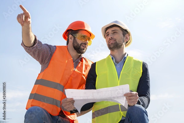 Fototapeta two men are builders workers are engineers at a construction site are looking at the drawings in helmets