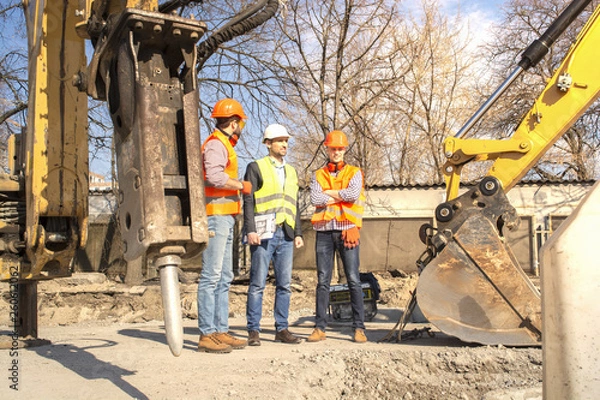 Fototapeta male workers engineers in helmets talking near the bulldozer and excavator