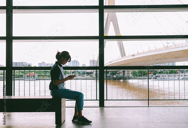 Fototapeta Asian woman sitting and used smartphone reads message- text message- take photos at the river scene background with copy space- education and business concept