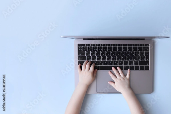 Fototapeta Kid hands typing on laptop keyboard on blue background, top view