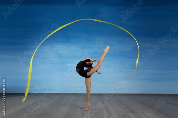 Obraz Girl gymnast in a black gymnastic swimsuit makes a circle in the air with a yellow ribbon on a blue sky background.