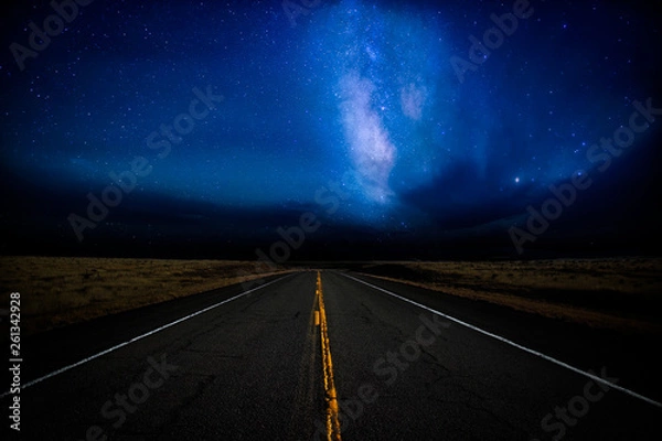 Fototapeta A highway disappearing into the distance illuminated by a star filled dramatic night sky in a rural landscape
