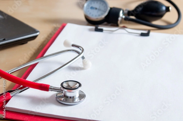 Fototapeta Red stethoscope on a red clipboard. Near laptop and tonometer. Copy space. Medical device. Treatment, health care. Heart examination. Studying the pulse. Close-up.