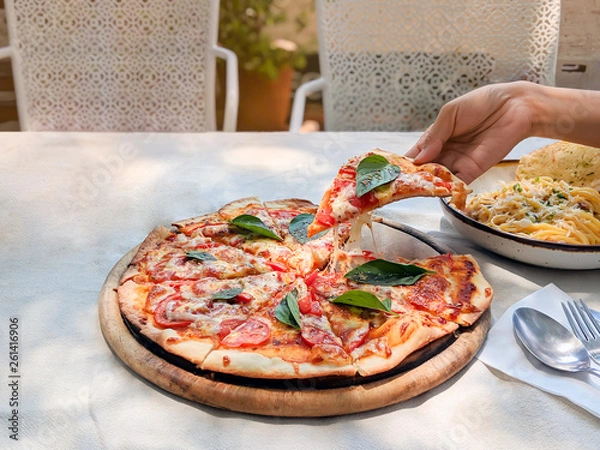 Obraz Hand is holding and pulling a slice of pizza margherita out of the tray,happy lunch time with friend in small garden. Foods on white tablecloth.