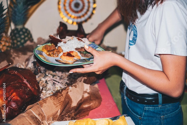 Fototapeta Young lady holding a plate of Filipino food while slicing a piece of meat of the pork roast or also known as Lechon  de leche.