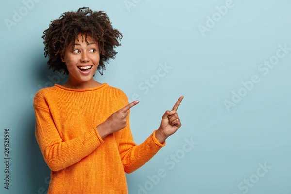 Fototapeta Poziome ujęcie zadowolona ciemnoskóra kobieta z fryzurą w stylu afro, wskazująca obiema przednimi palcami, pokazuje puste miejsce na promocję, odizolowane na niebieskim tle. Ludzie, koncepcja reklamy