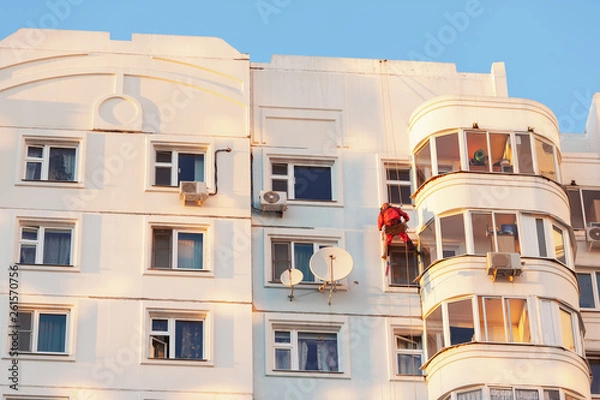 Fototapeta Industrial climber fixed on safety ropes puttying and painting the facade of building on sunny day. Bottom view from afar.