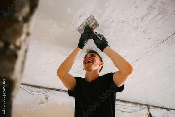 Fototapeta A man removes old paint from the ceiling with a spatula