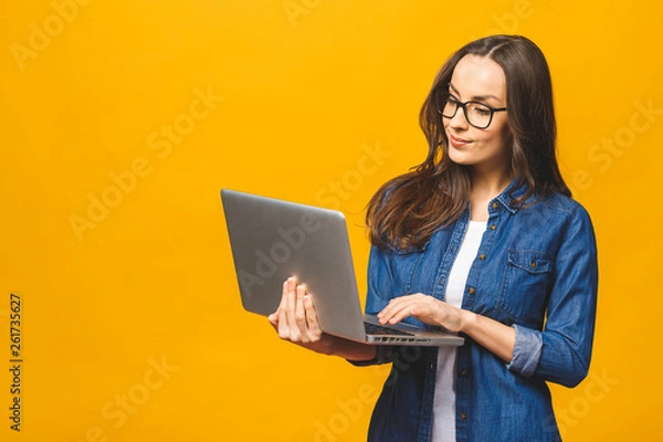 Fototapeta Młoda szczęśliwa uśmiechnięta kobieta w ubranie trzyma laptopa i wysyła e-mail do swojego najlepszego przyjaciela. Pojedynczo na żółtym tle.