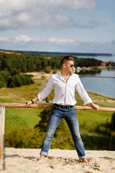 Fototapeta stylish guy in a white shirt is standing in the sand against the background of Nida