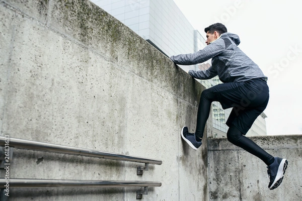 Obraz Stylish sportsman use architecture of urban area for his workout. He climbs at concrete wall, in the background modern office buildings.