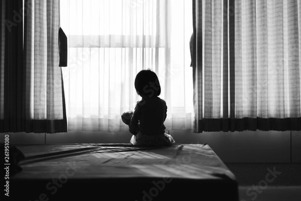 Fototapeta Little baby child sitting on bed playing with the doll in the dark room with light throught curtain