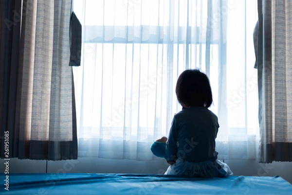 Fototapeta Little baby child sitting on bed playing with the doll in the dark room with light throught curtain