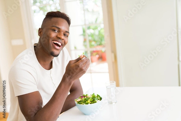 Fototapeta Handsome african young man eating a healthy vegetable salad using a fork to eat lettuce, happy and smiling sitting on the table