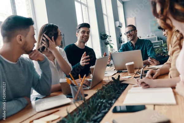 Fototapeta Achieving success together. Group of young modern people in smart casual wear discussing something and smiling while working in the creative office