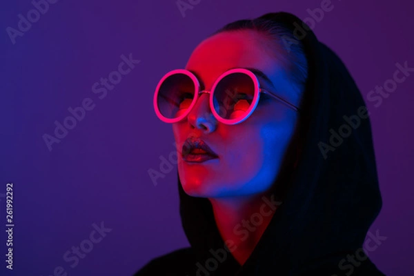 Fototapeta Portrait of fashion young girl in a black sweater with a hood and round sunglasses in red and blue neon light in the studio