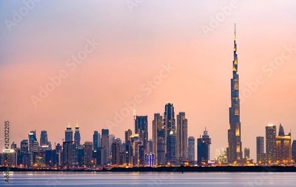 Obraz Oszałamiający widok na oświetloną panoramę Dubaju podczas zachodu słońca ze wspaniałym Burdż Chalifa oraz wieloma innymi budynkami i drapaczami chmur odbijającymi się w jedwabiście gładkiej wodzie płynącej na pierwszym planie.
