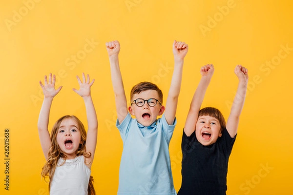 Fototapeta three children, a little boy and two little girls jumping up, their raised hands are happy, isolated yellow background, copy space