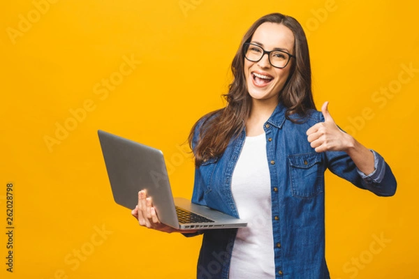 Fototapeta Portrait of a smiling young beautiful girl holding laptop computer and showing thumbs up isolated over yellow background.