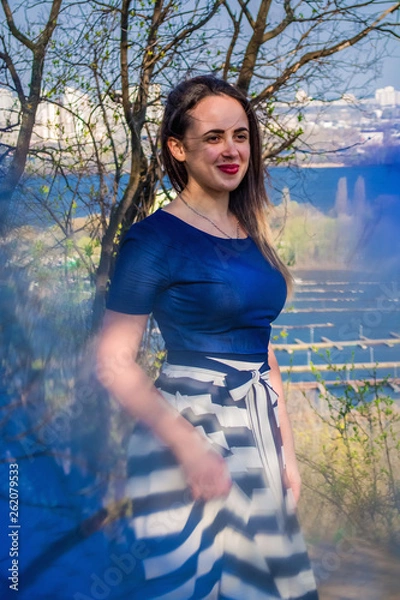 Fototapeta The young girl in a dress with a long skirt walks in the spring park. Blue a patch of light from a disk. Sunny weather. Fashion.