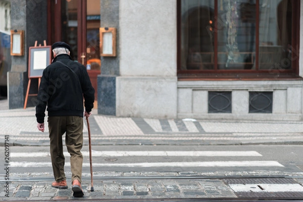 Fototapeta An elderly male with a walking stick slowly walks across a crosswalk in the middle of a city alone