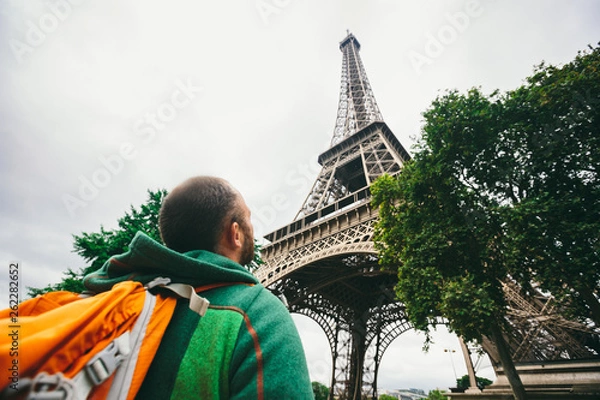 Fototapeta Caucasian male tourist with a backpack looking up and down at the top of the main attraction Paris Eiffel Tower in cloudy weather