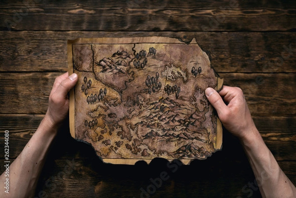 Fototapeta Old map in man adventurer hands on a brown table background. Treasure hunt concept.