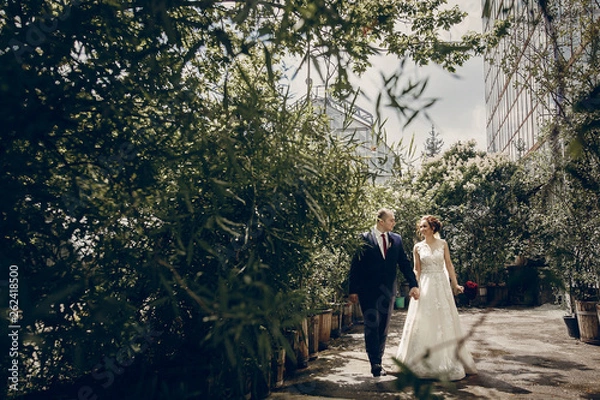 Fototapeta Romantic newlywed couple walking in the park, handsome groom and happy beautiful bride with bouquet holding hands in sunny botanic garden