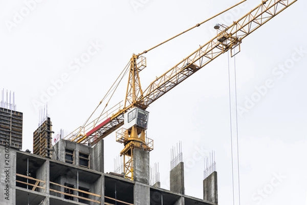 Fototapeta High-rise building crane with a long arrow of yellow color against the sky above a concrete building under construction with brick walls