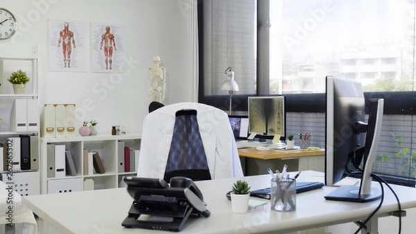 Obraz bright and modern medical doctor office complete with personal computer and telephone on desk white lab coat on chair. empty nobody in room in clinic. hospital workplace with sunshine through window.