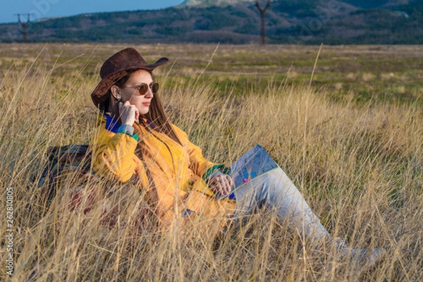Fototapeta A young girl with long dark hair in a yellow jacket, leather cowboy hat and glasses sits in the same grass, leaning on a tourist backpack, looking into the distance. Background mountains, sky.