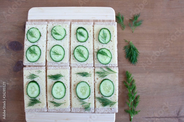 Fototapeta Light, spring sandwiches with cheese, fresh cucumber and dill, next to a tray with slices of cucumber and a board full of green spring vegetables