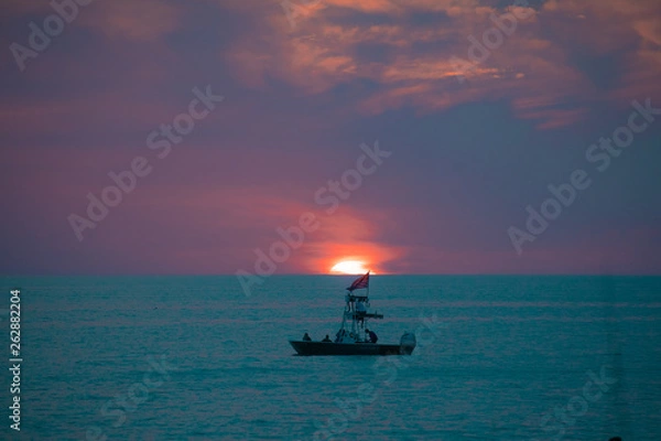 Fototapeta A boat on the teal green ocean water in front of the setting sun amid the blue and purple clouds of evening, as seen from a beach on the Gulf of Mexico near Englewood, Florida, USA, in early spring