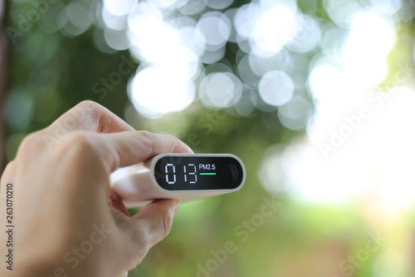 Obraz measuring outdoors air quality. A hand holding particulate matter 2.5 (pm.2.5) sensor. harmful small dust detector indicated healthy acceptable air quality. blur background of blue green plants.